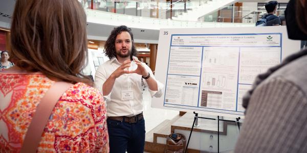 Man with long hair gestures while describing research poster to audience members