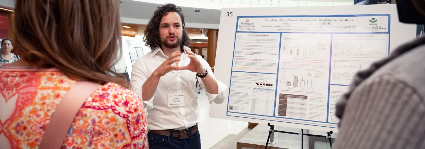 Man with long hair gestures while describing research poster to audience members